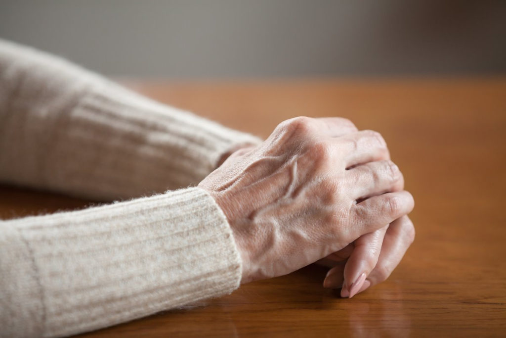 Close up view of mature old female person wrinkled hands with veins on table, middle aged senior elderly woman holding arms folded praying as concept of aging process, healthcare, loneliness or grief